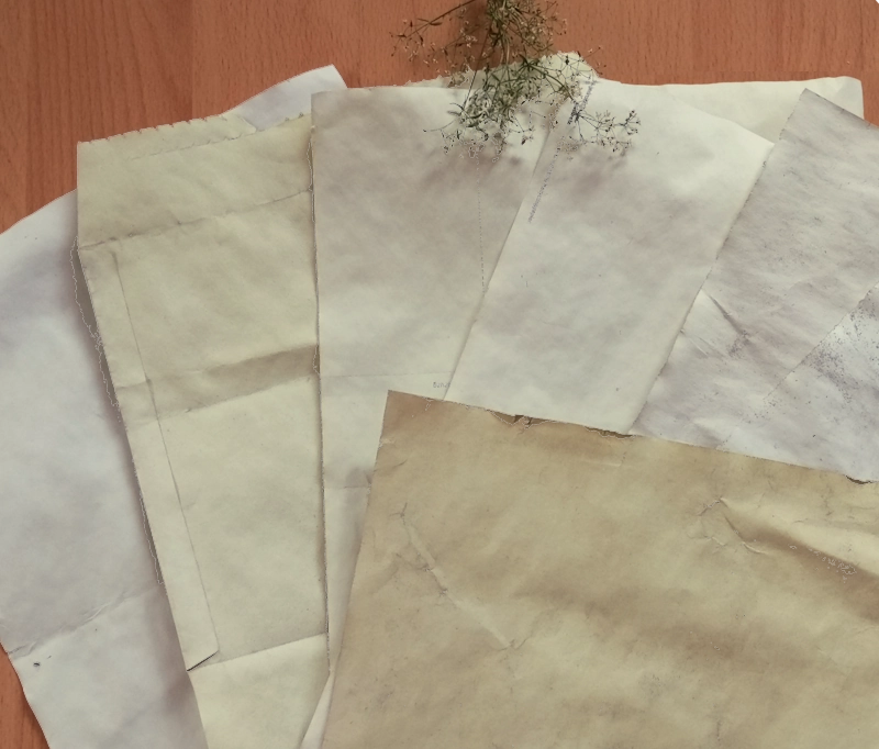 Papier färben mit Naturmaterialien: Papier in verschiedenen Grüntönen, die mit Brennnesseln gefärbt wurden. Der Hintergrund ist aus Holz und zur Dekoration ist ein kleiner Blumenstrauß am oberen Rand zu sehen.