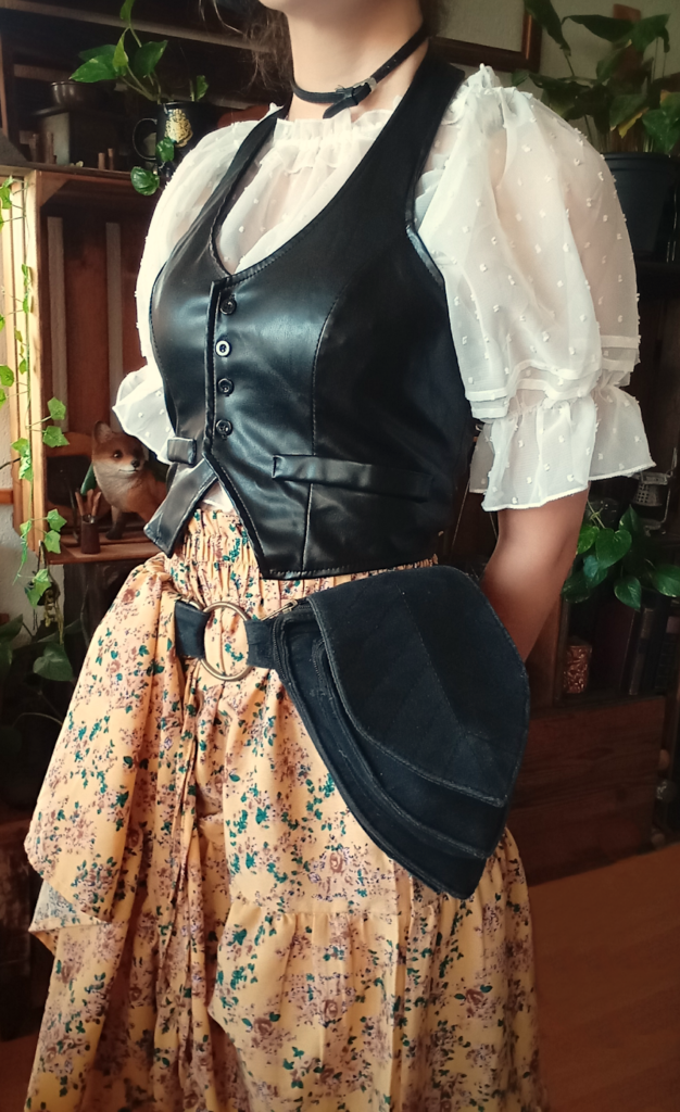 Outfit mit gelbem, geblümelten, langem Rock, schwazer Bauchtasche in Blatt-Form, weißer Bluse mit Puffärmen und einer schwarzen Weste - im Hintergrund sind Holzkisten und Pflanzen.