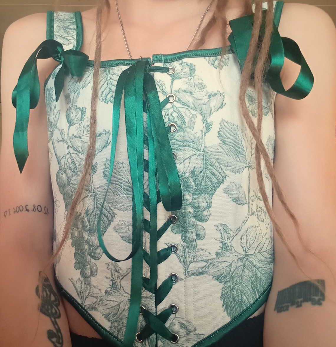 Nahaufnahme eines getragenen, weißen Korsets. Grüne Aufdrucke von Trauben - 3 Tattoos auf den Armen