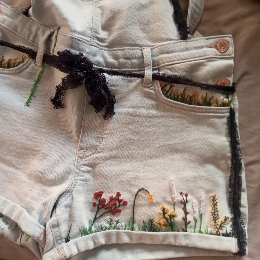 Stickarbeit einer Blumenwiese und von gesticktem Farn auf der Hinterseite eines Overalls. Schwarze Spitze ist an den Overall genäht.