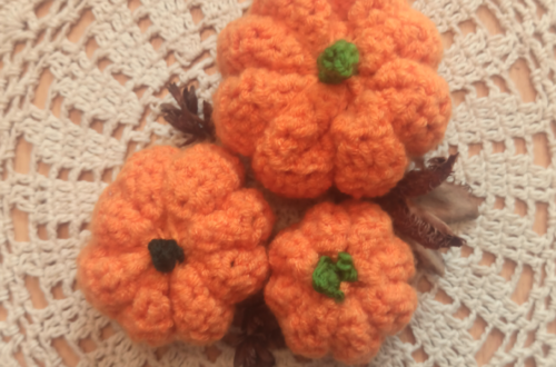 3 gehäkelte, orangefarbene Kürbisse auf einem weißen Zierdecken auf Holzuntergrund